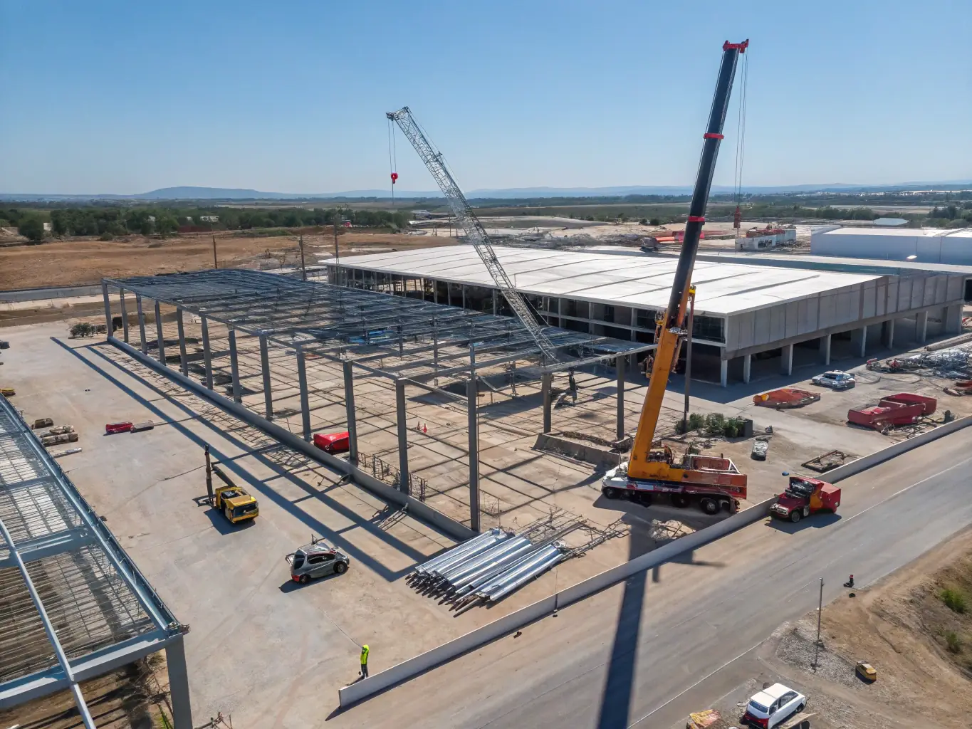 A wide shot of a large industrial warehouse under construction, featuring steel framework and heavy machinery. Construction workers are visible, wearing safety gear and operating equipment.